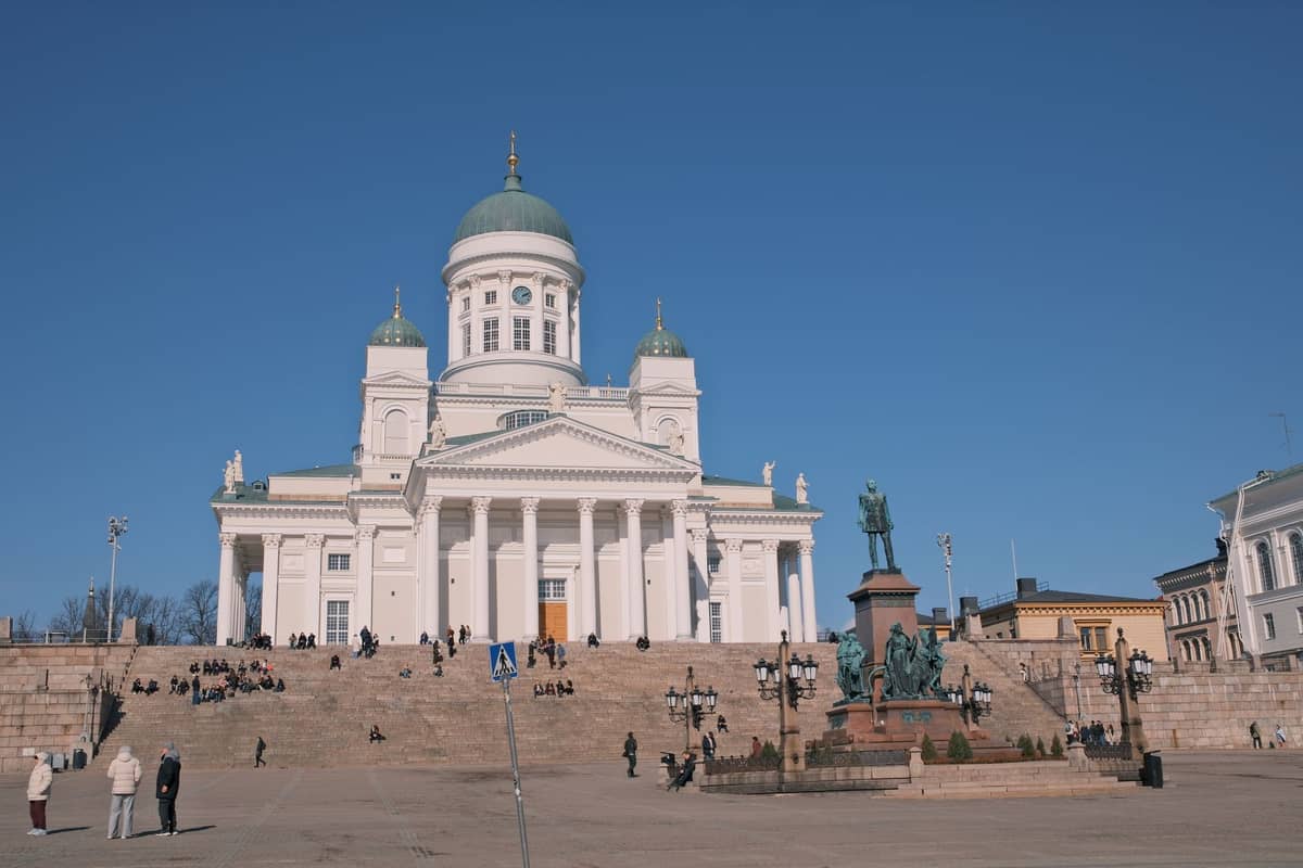 Vista ampla da Catedral de Helsinque e da Praça do Senado com pessoas caminhando e se reunindo na área aberta sob um céu azul claro.