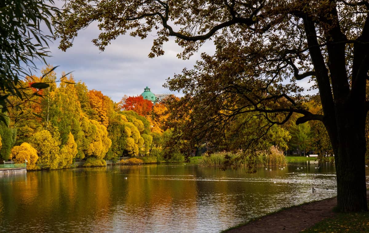 Vista sobre um lago em um parque da cidade de Helsinki com árvores de outono vibrantes, reflexos na água e um edifício com cúpula ao fundo.