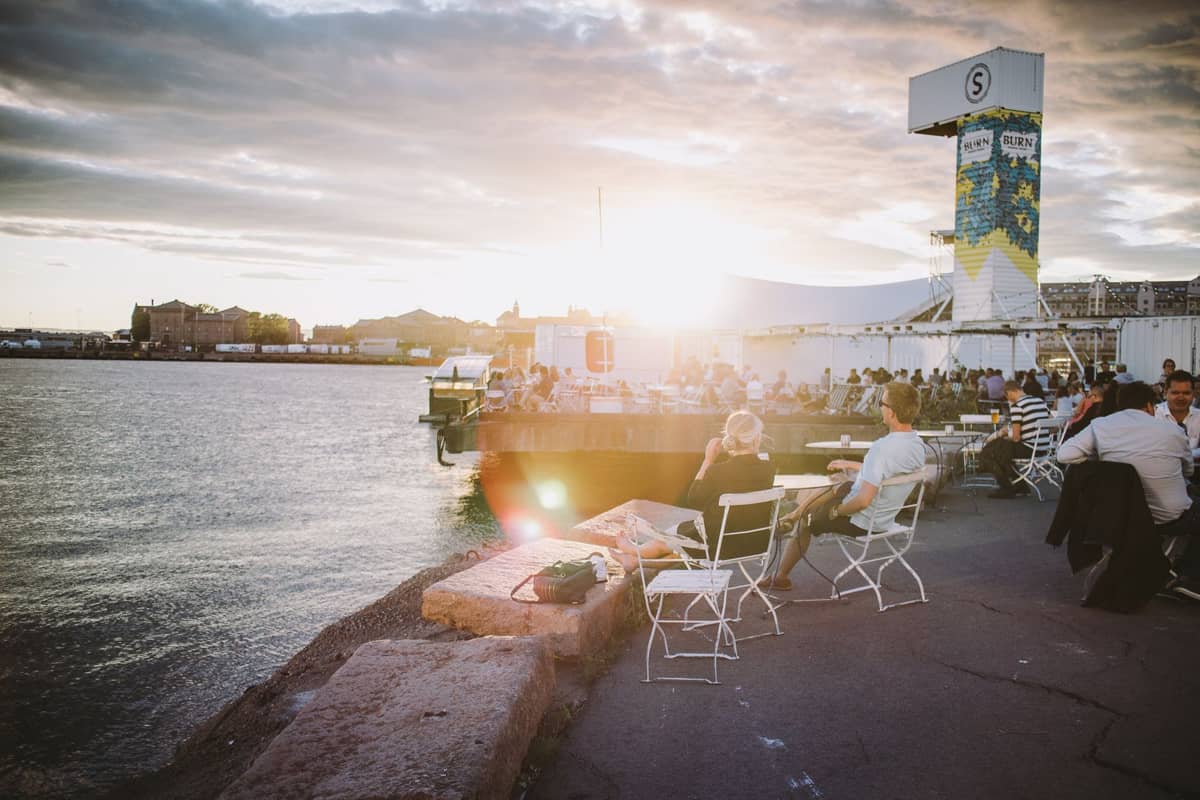 Pessoas relaxando em assentos ao ar livre à beira-mar com o brilhante sol de verão sobre o porto de Helsinque, atmosfera animada e vibrante clima de festival moderno.