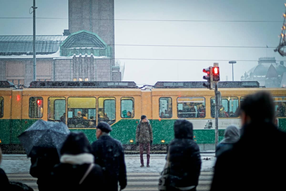 Neve cai sobre pessoas que esperam numa passadeira perto de um elétrico amarelo e verde em frente à Estação Central de Helsínquia.