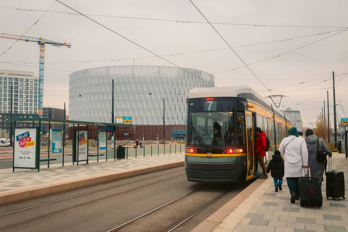 Bonde moderno de Helsinque em uma parada da cidade, com adultos e crianças embarcando, malas e carrinho de bebê à vista, destacando o transporte público para famílias.