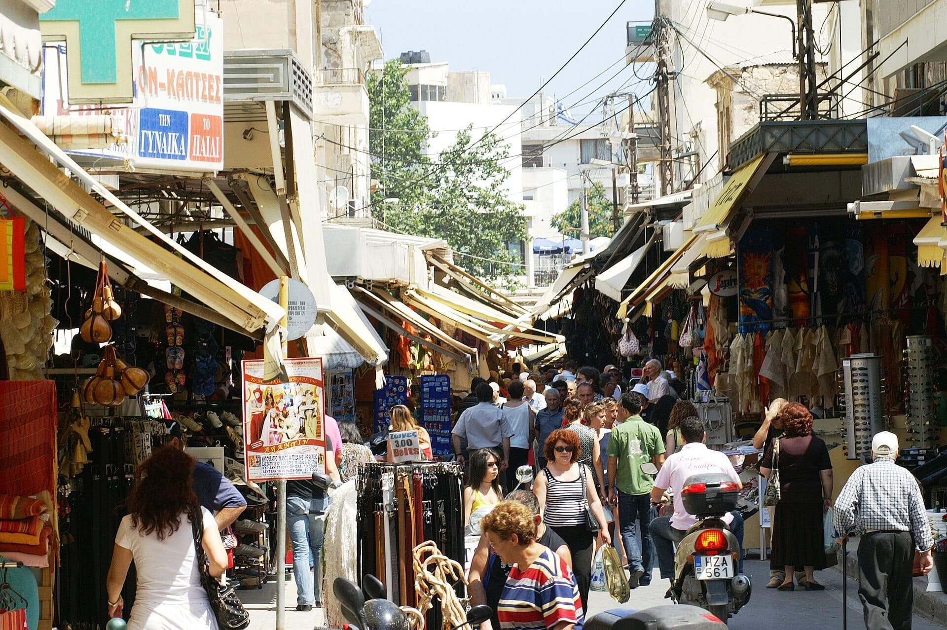 Vivace scena diurna al mercato di Via 1866 a Heraklion, con persone che camminano tra negozi aperti, tettoie e bancarelle colorate.