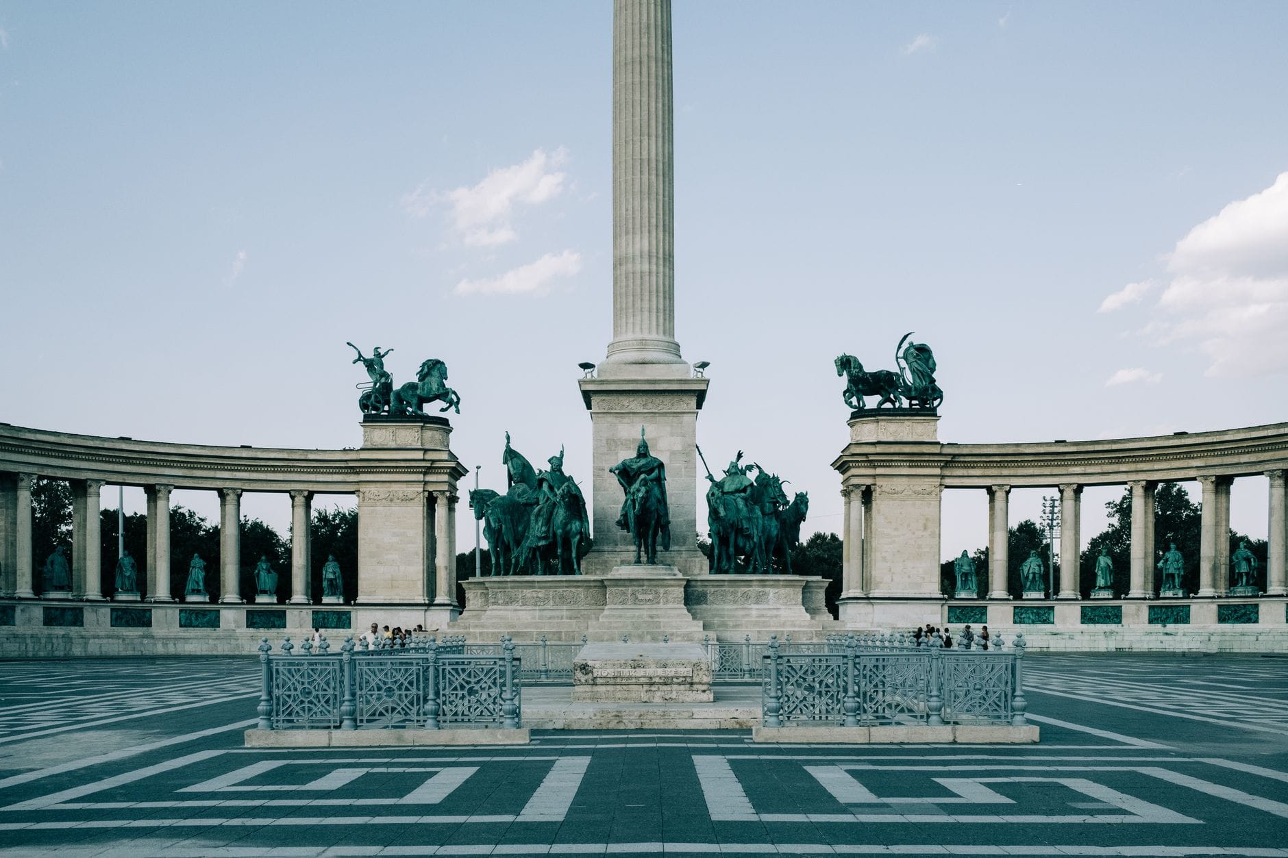 Veduta diurna di Piazza degli Eroi a Budapest, con il Monumento del Millennio, le statue circostanti e la grande piazza aperta sotto un cielo sereno.