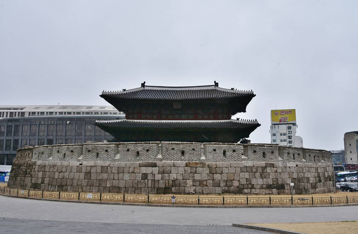 O Portão Heunginjimun (Portão Dongdaemun) em Seul, mostrando o histórico portão de pedra e suas muralhas circulares, com prédios da cidade ao fundo em um dia nublado.