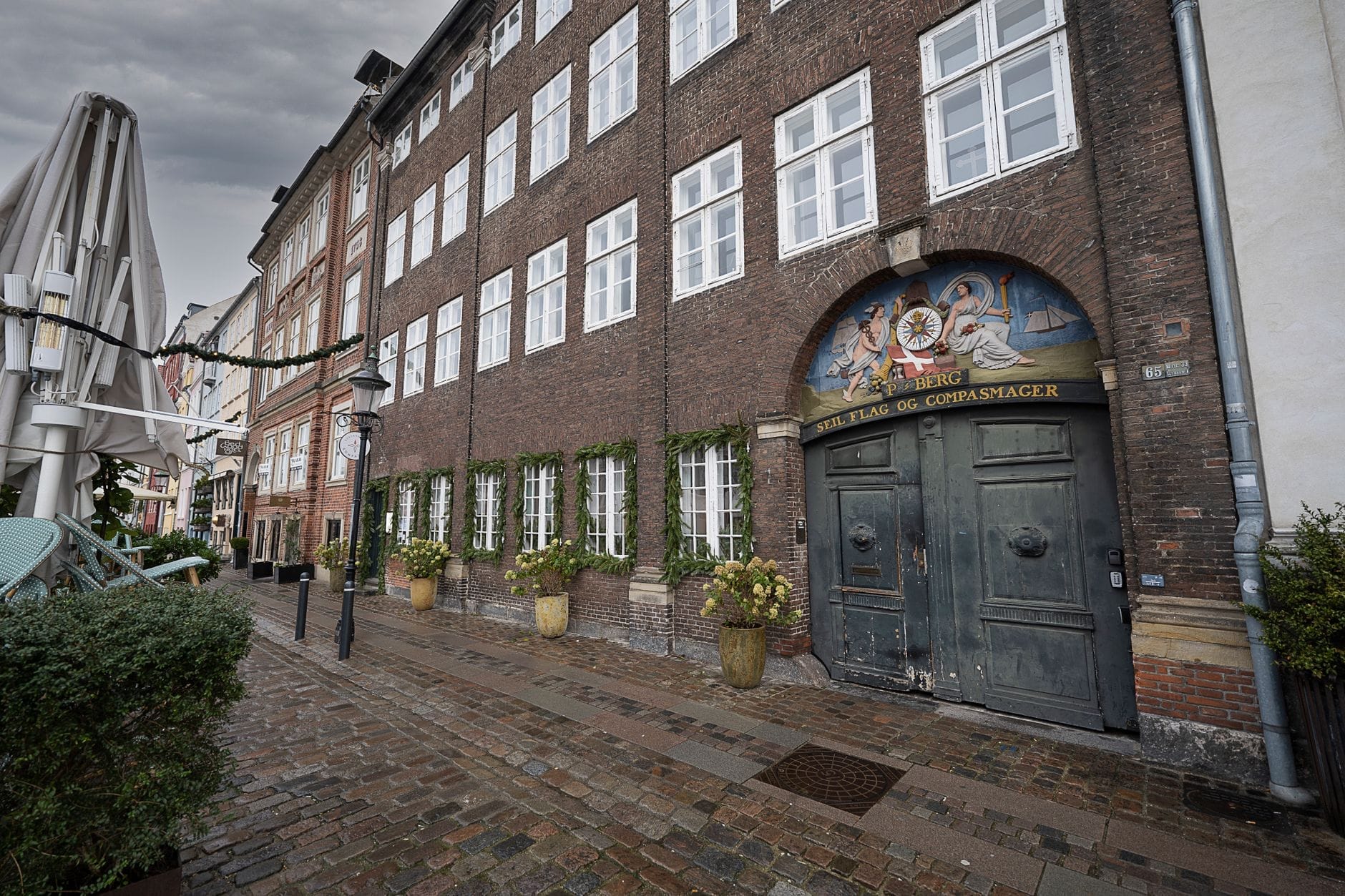 Uma rua de paralelepípedos em Copenhague ladeada por prédios antigos de tijolo, grandes janelas e vasos de plantas verdes, sob um céu nublado e sem multidões.