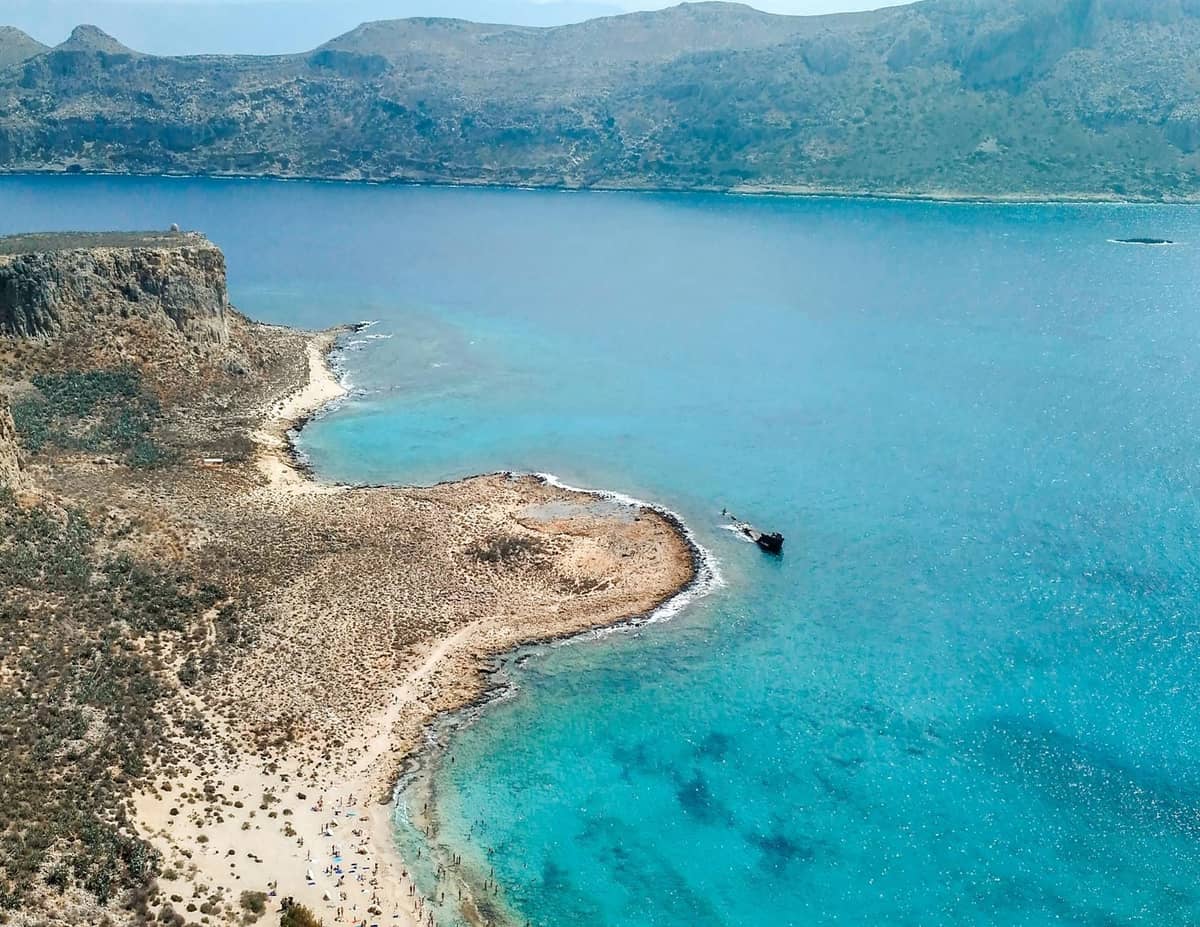 มุมมองทางอากาศของชายหาดที่ห่างไกลและขรุขระบน Crete พร้อมน้ำสีฟ้าเทอร์ควอยซ์ ชายฝั่งทรายสวยงาม และเนินเขาที่เป็นหินผา สะท้อนให้เห็นธรรมชาติที่ยังคงความบริสุทธิ์