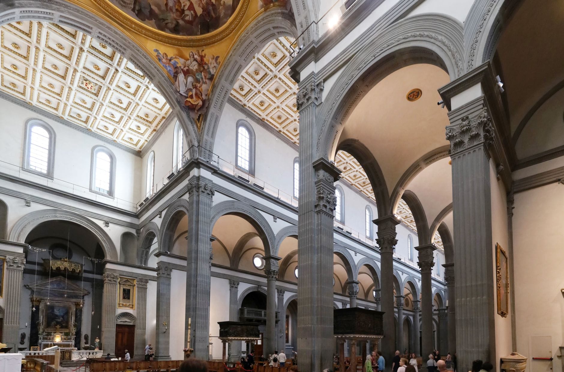 ภายในโบสถ์สไตล์เรเนซองส์ในเมือง Florence ที่มีเสาหินสีเทา เพดานลายกรอบสี่เหลี่ยม โค้งประตู และผู้คนที่กำลังชื่นชมสถาปัตยกรรม