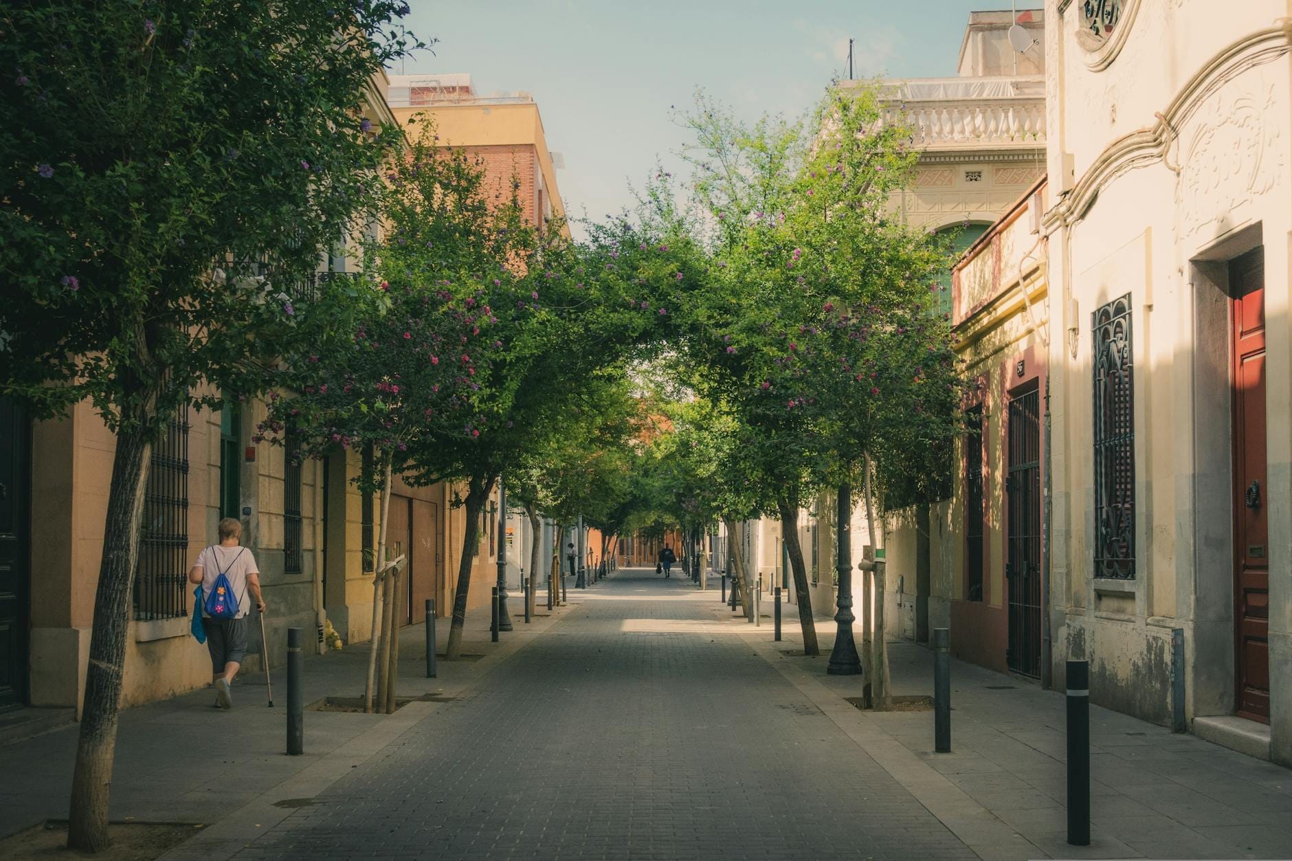 A peaceful, sunlit Barcelona street with low-rise buildings, arching green trees, and a few people strolling down the shaded pedestrian walkway.