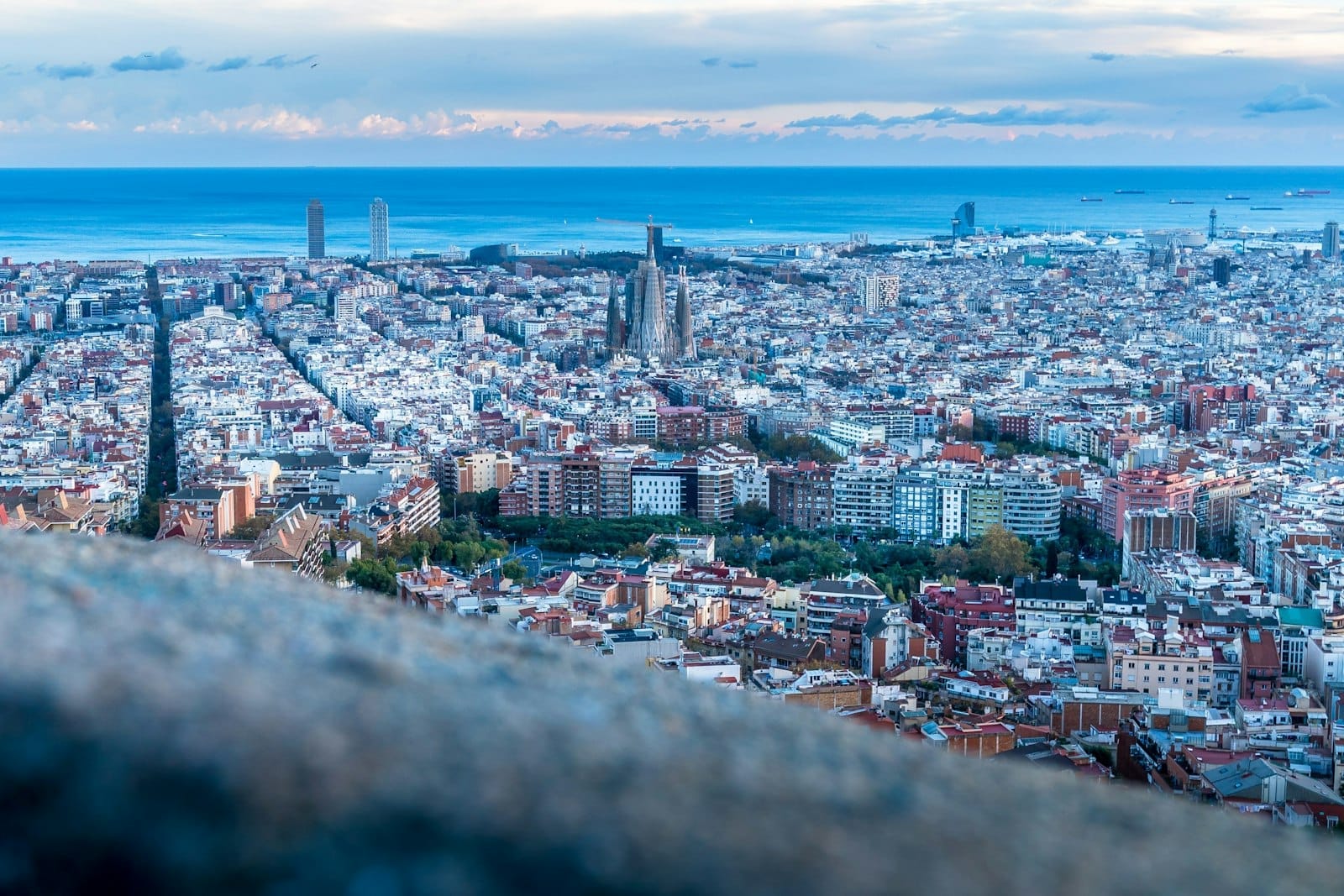 A wide aerial view of Barcelona at dusk, featuring the iconic Sagrada Família, grid-like streets, colorful buildings, and the Mediterranean Sea in the distance.