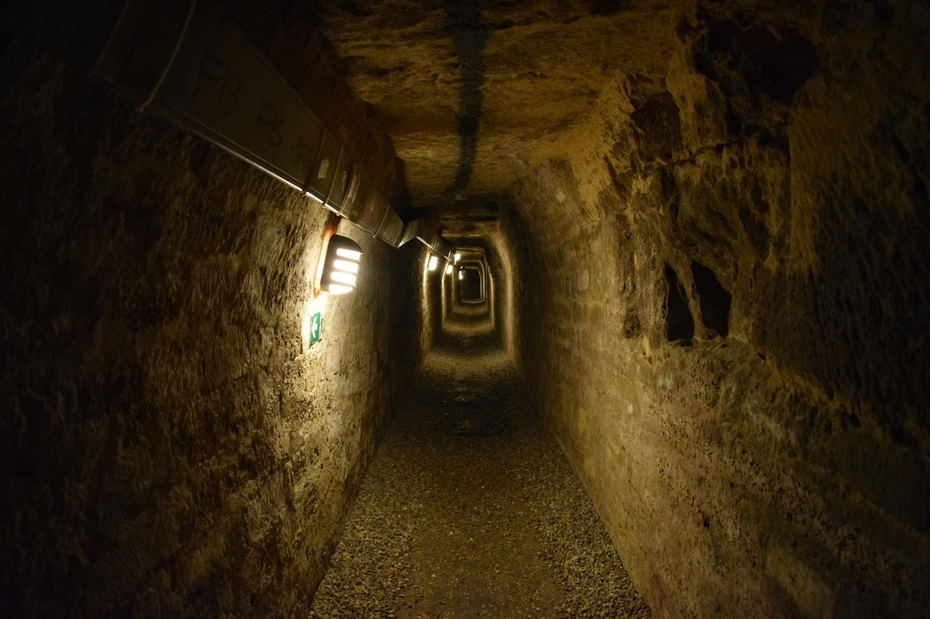 Schwach beleuchteter Steintunnel mit rauen Wänden und Decke, gesäumt von Lampen, der eine atmosphärische unterirdische Szene erzeugt.