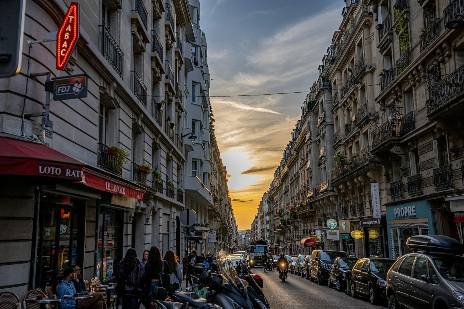 Una vivace strada parigina al tramonto con passanti, scooter e palazzi classici su entrambi i lati sotto un cielo serale spettacolare.