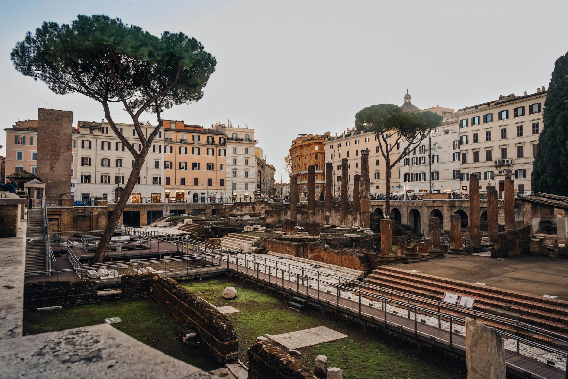 View of ancient Roman ruins with columns and underground excavations, surrounded by typical Roman buildings in central Rome.