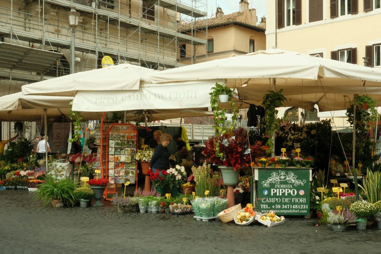 Flower and produce market stall under white canopy in a Roman piazza with fresh plants, a vendor, and historic buildings behind.