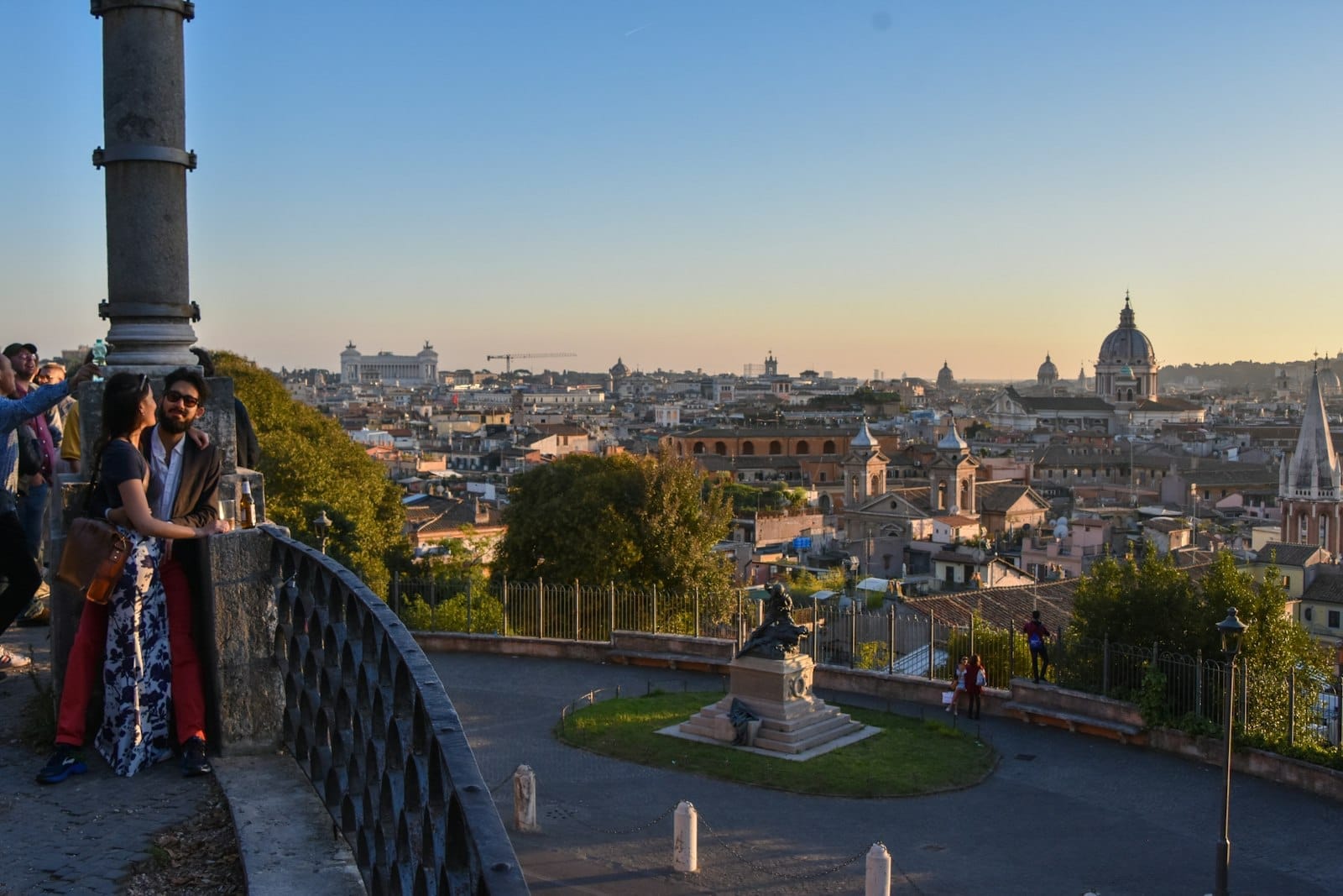 People enjoying a scenic city panorama of Rome at sunset from a hilltop terrace with domes and rooftops visible.