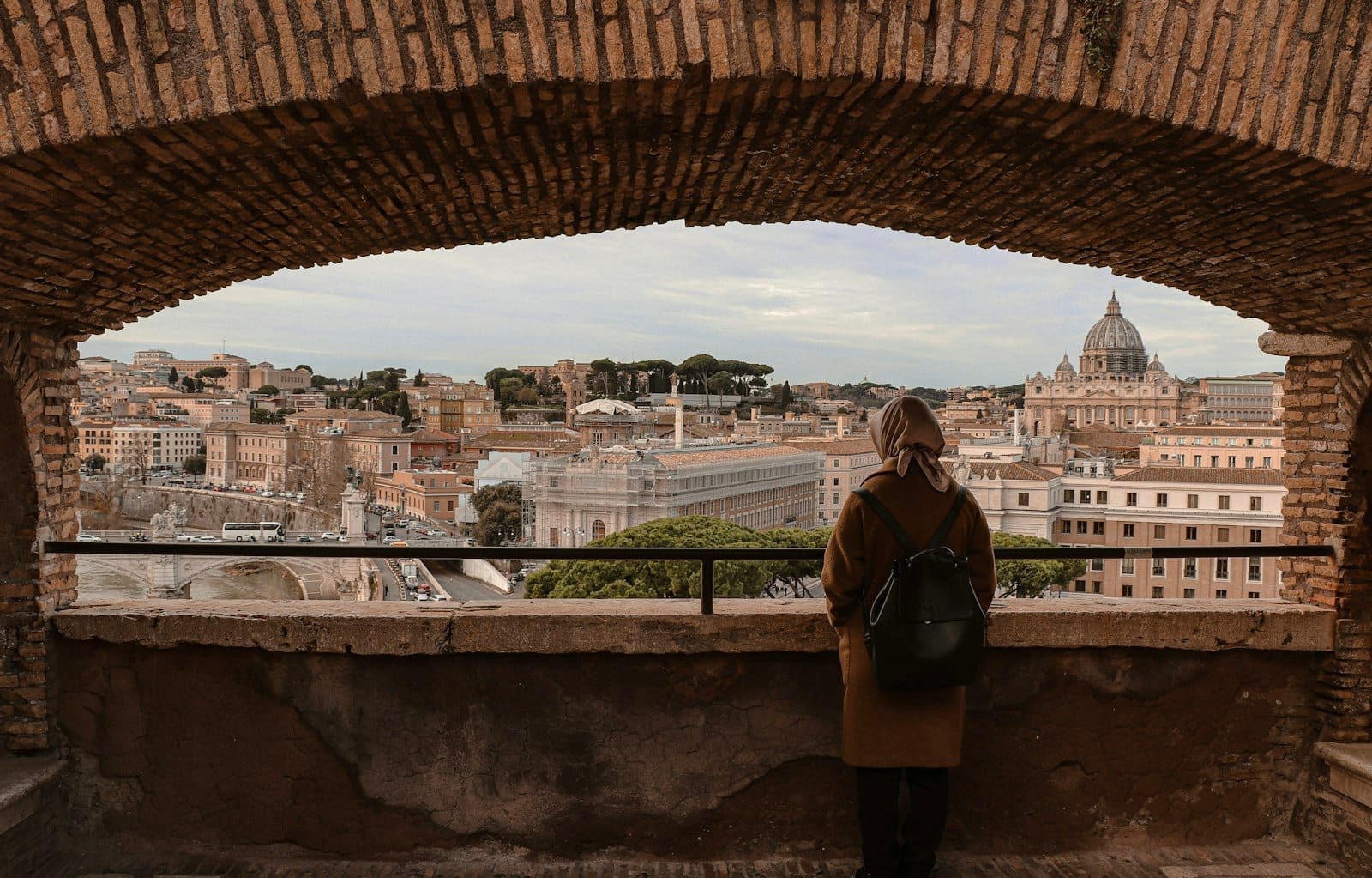 A person with a backpack looks out over rooftops of Rome through a stone archway, with St. Peter’s Basilica visible in the distance.