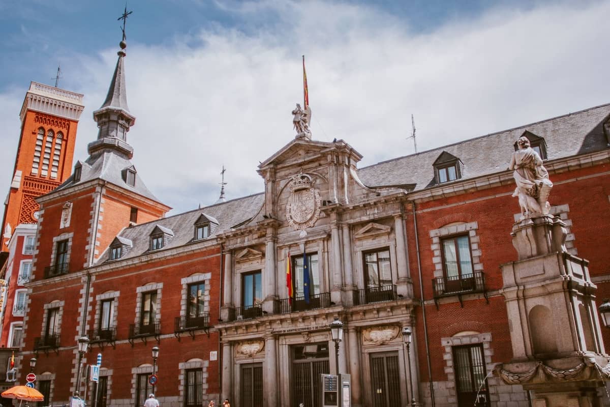Historisches Backsteingebäude in Madrid mit kunstvollen architektonischen Details und einer Statue davor unter einem teils bewölkten Himmel.