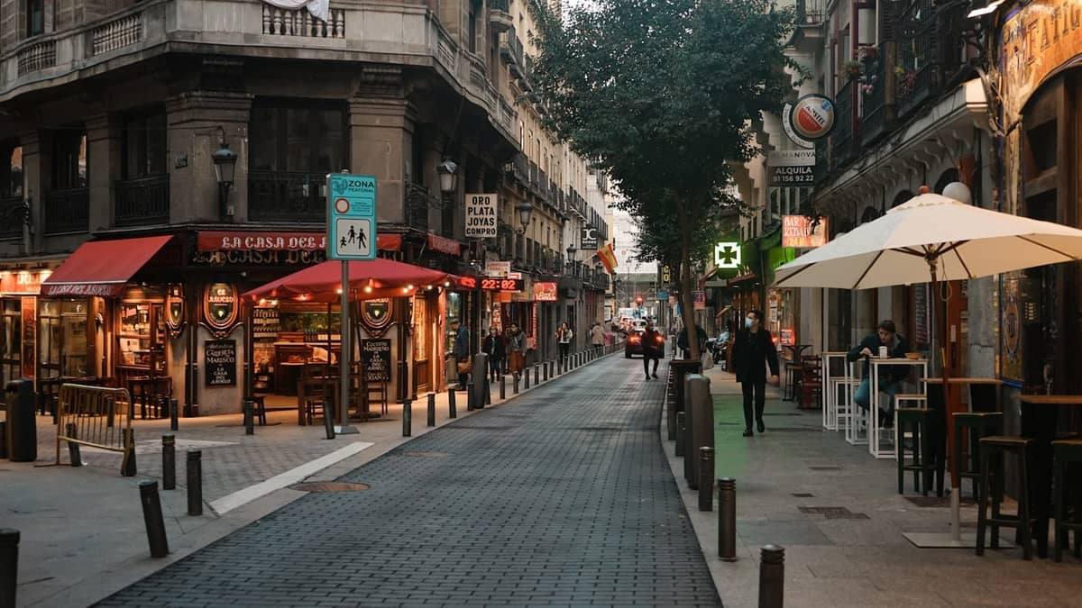 Eine lebhafte Straße in Madrid, gesäumt von Cafés und Restaurants, mit Außensitzplätzen, Fußgängern und klassischer Architektur, die die lokale Gastronomieszene eines Viertels perfekt einfängt.