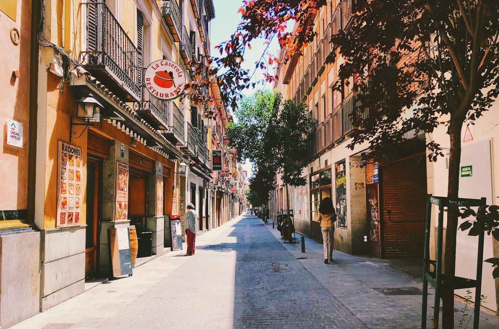 Rua lateral ensolarada e tranquila em Madri com prédios coloridos, árvores que fazem sombra, placas de café ao ar livre e uma única pessoa caminhando, convidando à exploração além das multidões turísticas.
