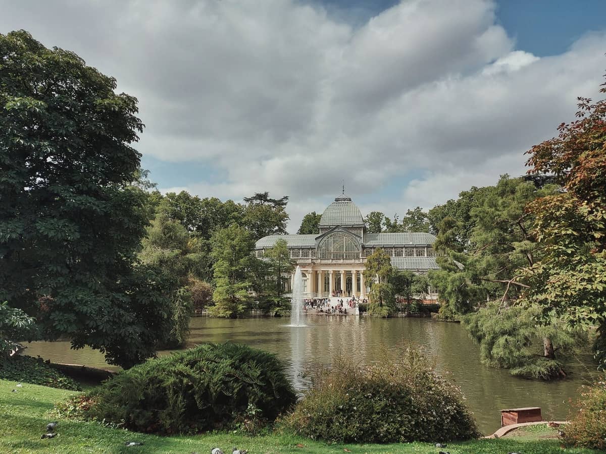Eine ruhige Gartenszene mit dem gläsernen Palacio de Cristal, einem Teich, umgebendem Grün und einem Brunnen im Parque del Retiro in Madrid.