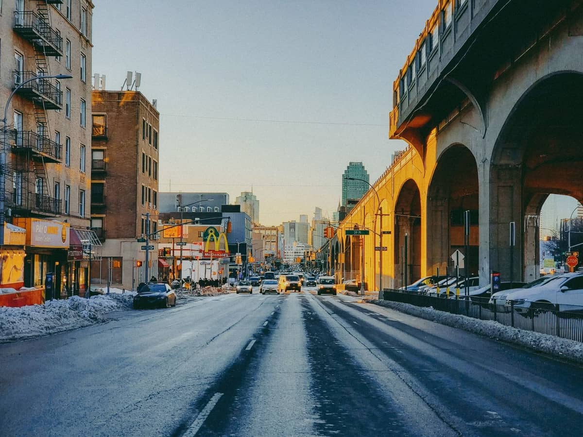 Städtische Straße in Queens oder Brooklyn mit erhöhten U-Bahn-Gleisen, Vintage-Gebäuden und fernem Stadtpanorama im goldenen Winterlicht.