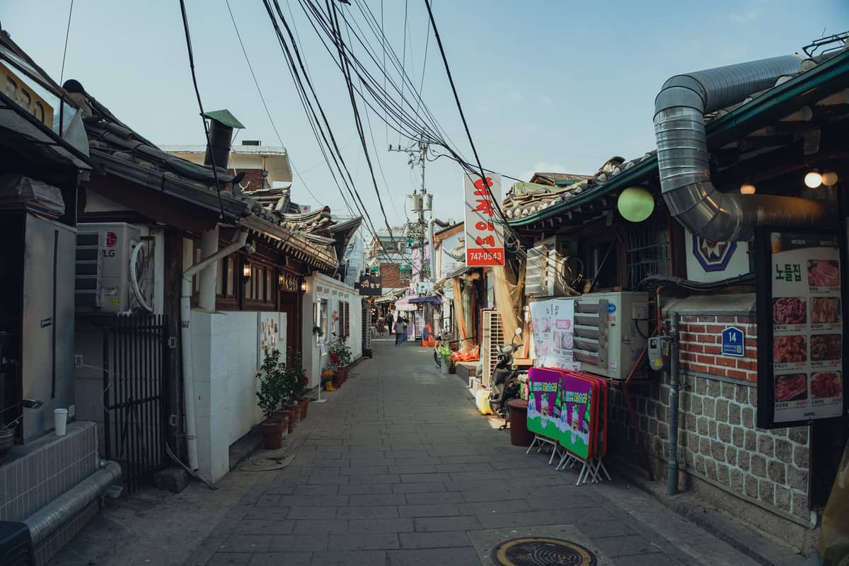 Um beco tradicional estreito em um bairro pouco conhecido de Seul, ladeado por casas no estilo hanok, fios suspensos, placas de lojas e restaurantes locais sob um céu limpo.
