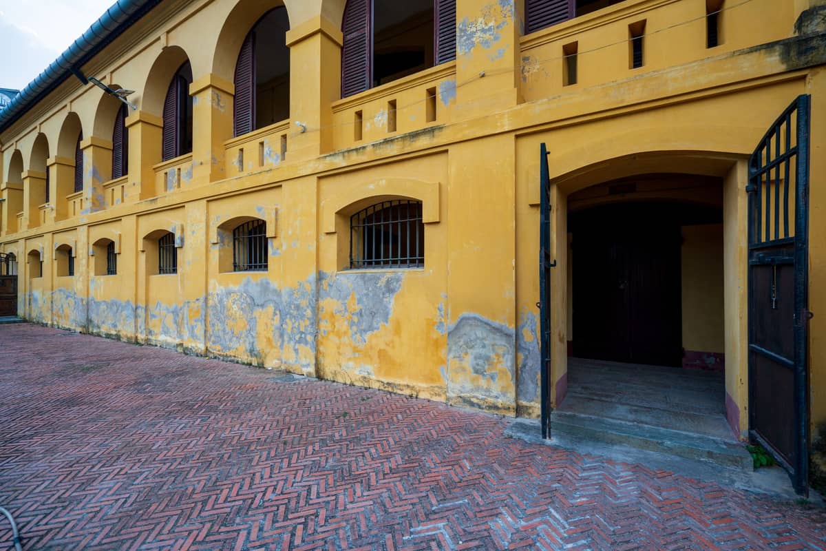 Edifício de concreto amarelo e envelhecido com janelas gradeadas e um portão de ferro aberto que leva ao interior, evocando uma prisão histórica ou um local institucional.