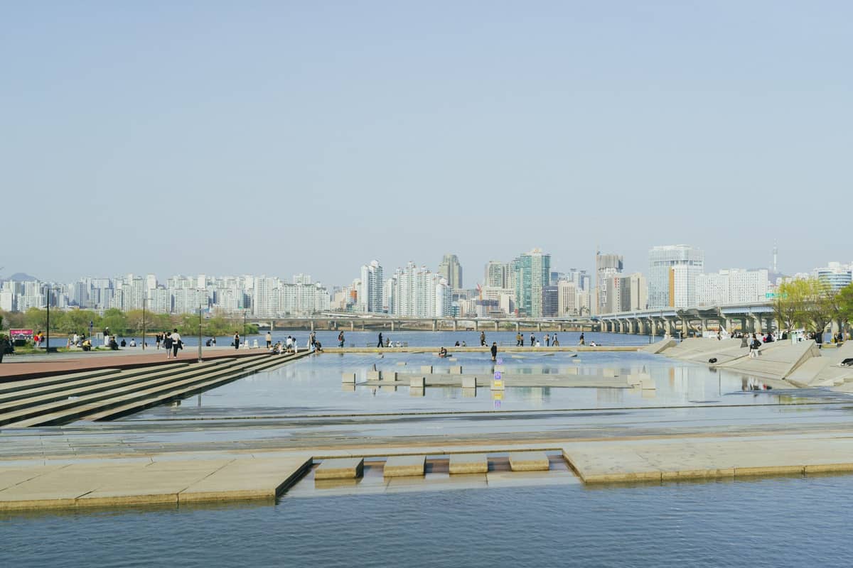 Vista ampla das terraças aquáticas do Parque Seonyudo com pessoas sentadas e caminhando, o Rio Han e o horizonte de Seul ao fundo em um dia ensolarado.