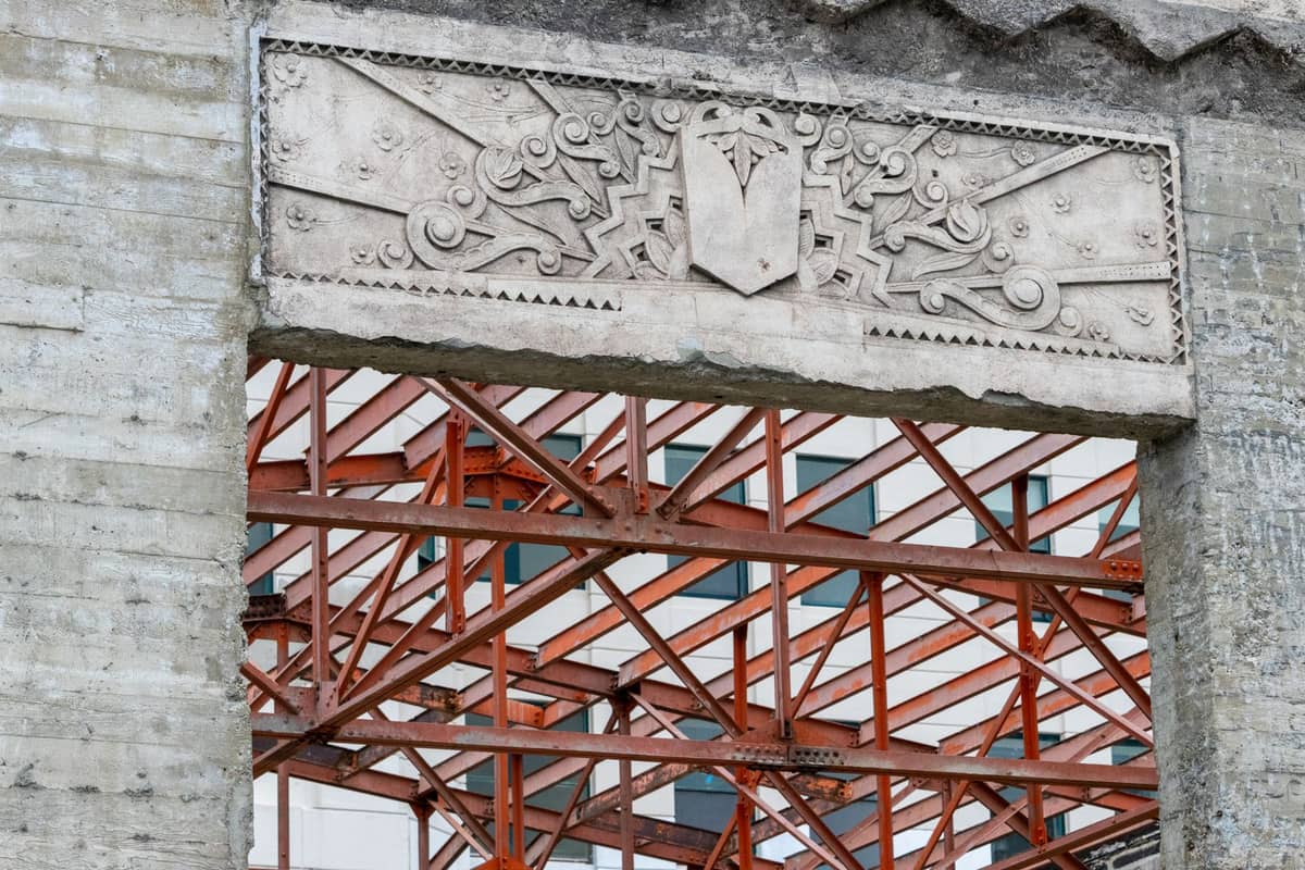 Detalle de una estructura de concreto antigua con relieve decorativo sobre un vano de puerta, mostrando vigas de acero naranja expuestas detrás