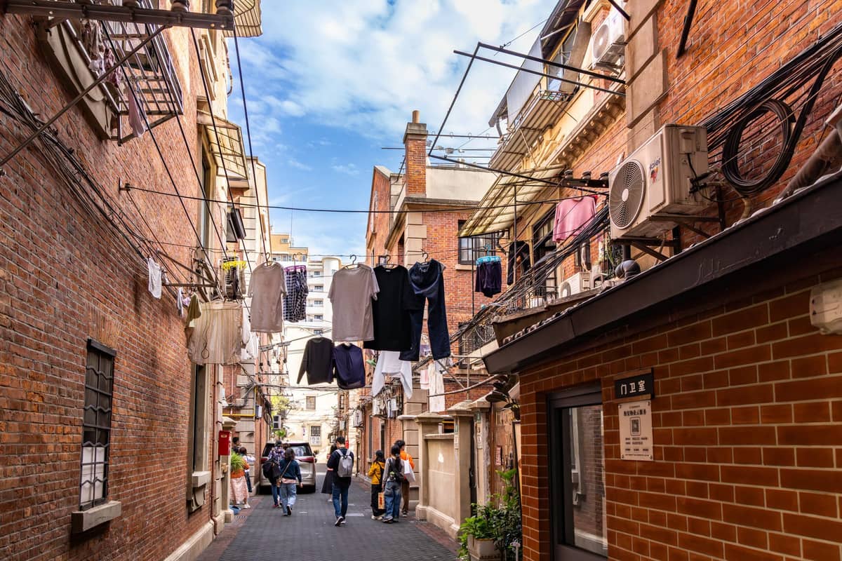 Enge Shanghaier Gasse mit rotbacksteinernen Shikumen-Häusern, Wäscheleinen über den Köpfen und Menschen unter blauem Himmel.