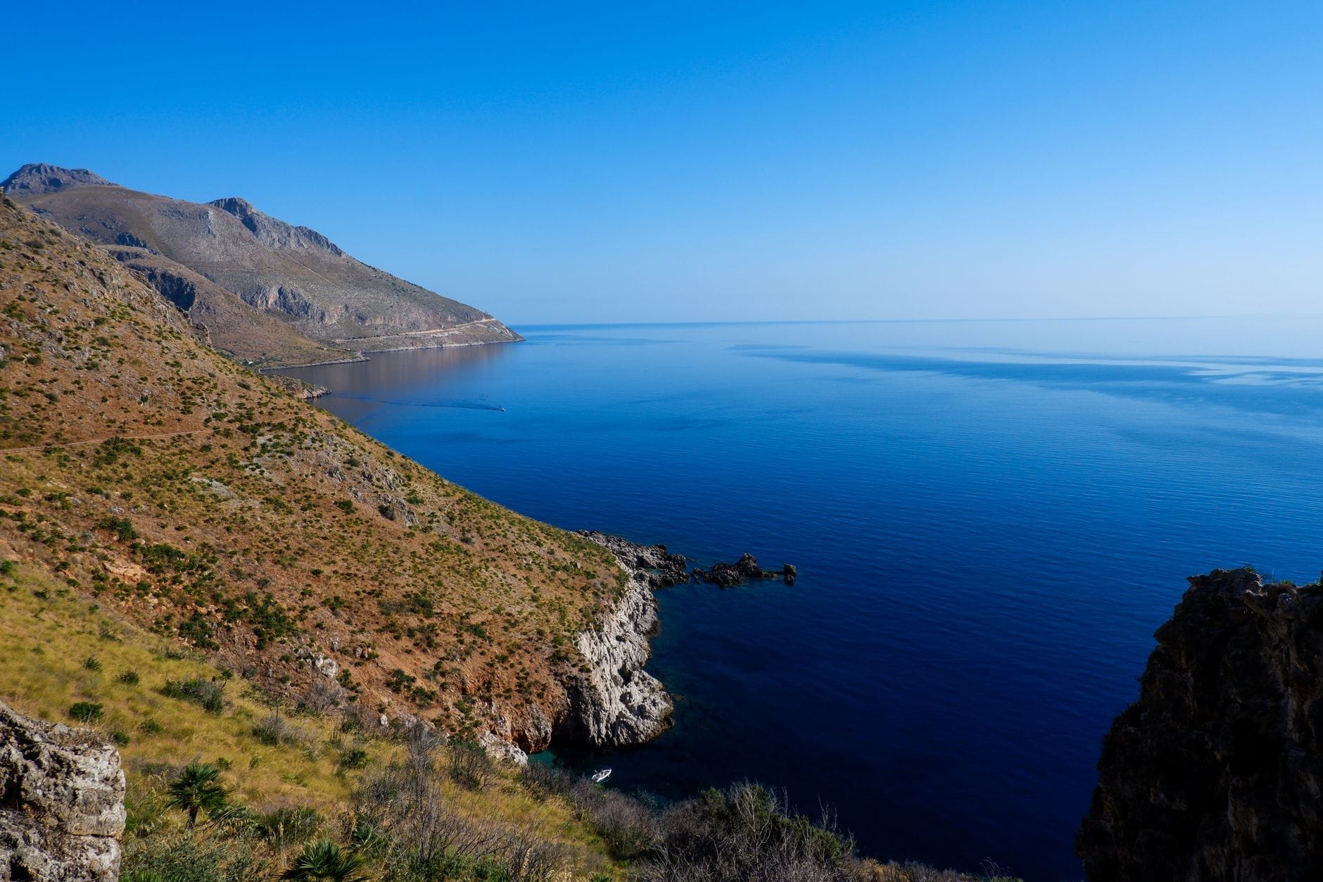 Küstennaturschutzgebiet mit schroffen Klippen, klarem blauem Meer und einer bergigen Kulisse unter einem hellen Himmel in Sizilien.