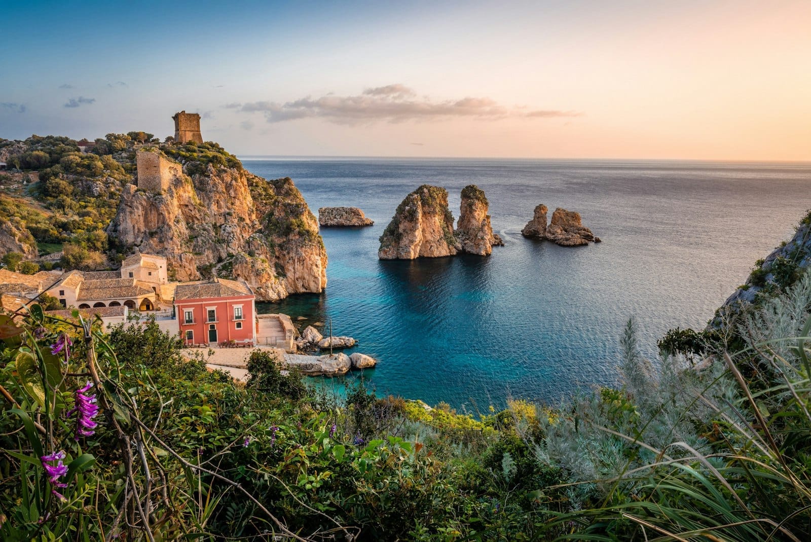 Dramatische Küstenfelsen mit einem historischen Wachturm, rustikalen Gebäuden, türkisfarbenem Meer und Felsstapeln bei Sonnenuntergang, umgeben von Wildblumen und üppiger mediterraner Vegetation in Sizilien.