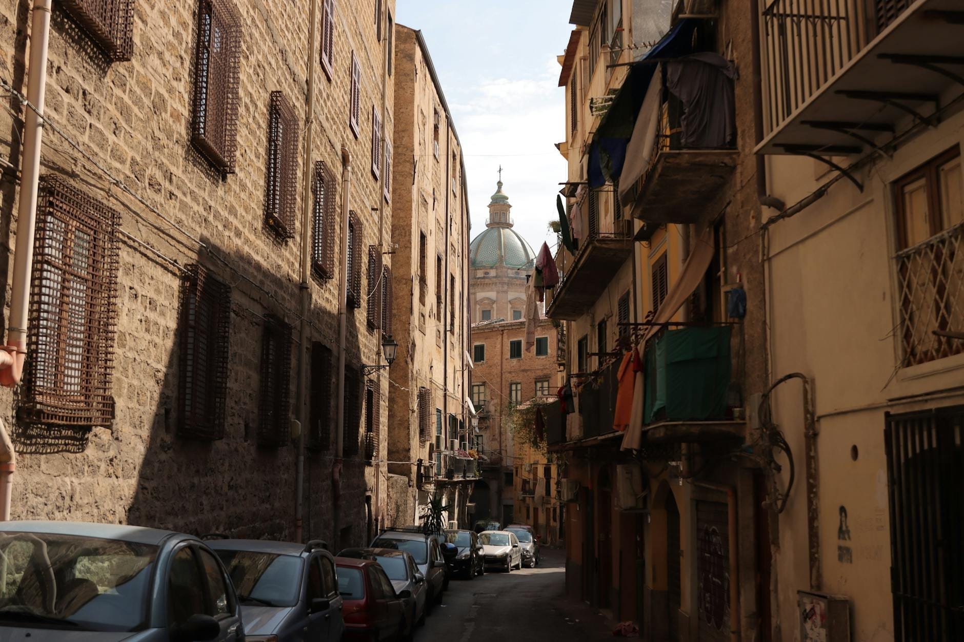Enge Straße in Palermo, gesäumt von alten Gebäuden, geparkten Autos, Balkonen und der Kuppel einer historischen Kirche in der Ferne.