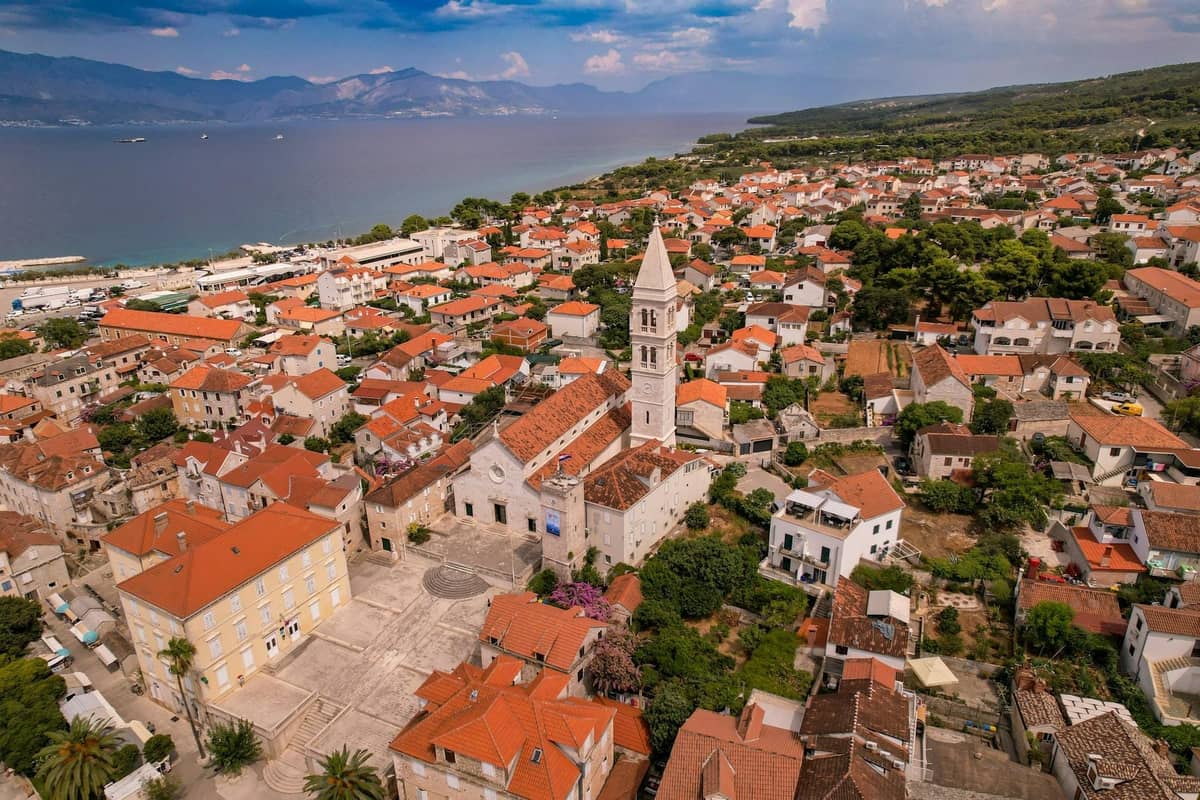 Luftaufnahme einer kleinen Küstenstadt in der Nähe von Split mit einer Kirche mit einem hohen Glockenturm, Häusern mit roten Dächern und dem blauen Meer im Hintergrund.
