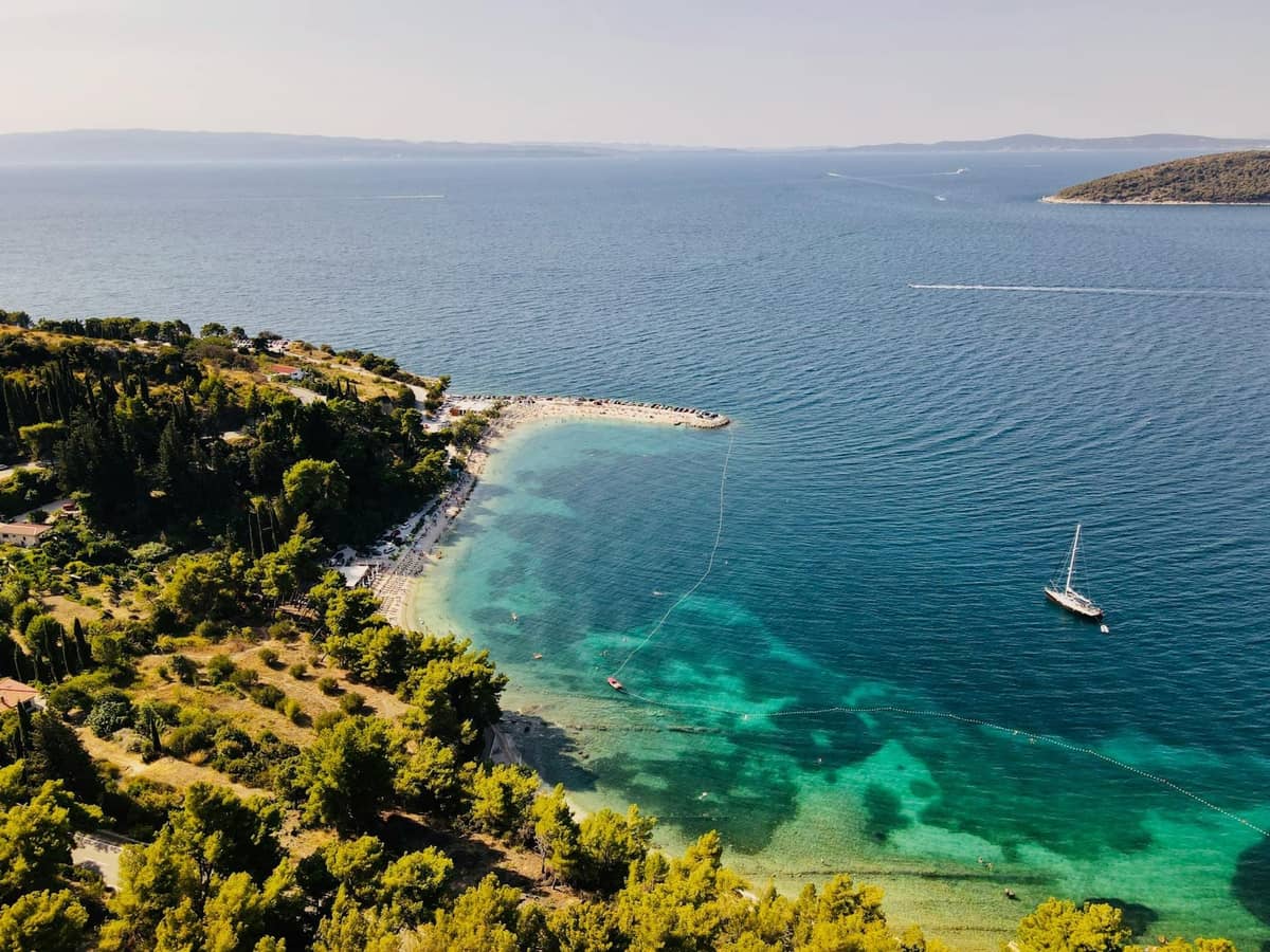 Luftaufnahme eines ruhigen Felsstrandes in der Nähe von Split mit türkisfarbenem Wasser, bewaldeter Küste und einer vor Anker liegenden Yacht unter klarem Himmel.