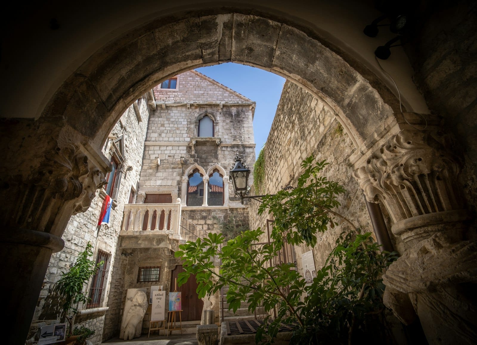 Pátio medieval de pedra em Split emoldurado por um arco antigo, com vegetação exuberante, colunas ornamentadas e luz do sol nas fachadas históricas.
