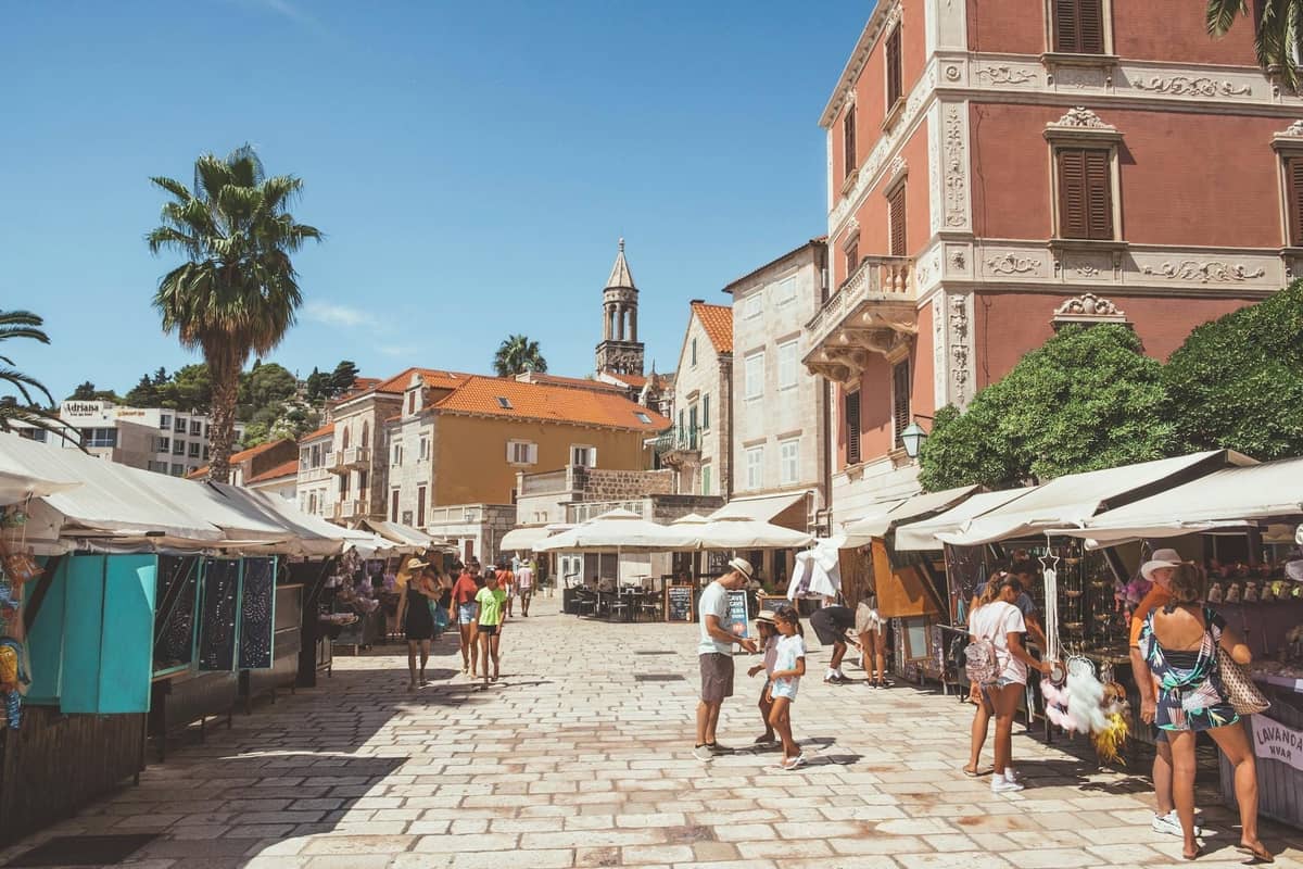 Outdoor-Marktszene in Split mit Menschen, die an Ständen auf einem gepflasterten Platz einkaufen, der von historischen mediterranen Gebäuden gesäumt wird.