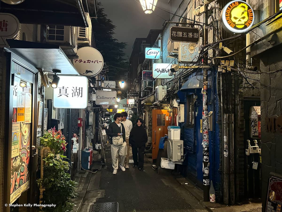 Narrow alley in Tokyo's Golden Gai at night with bright bar signs, glowing lanterns, and small groups of people walking between cozy bar entrances.