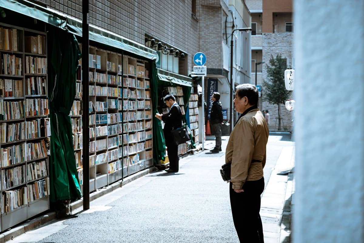 Outdoor shelves filled with books line a narrow Tokyo street, with several people browsing under daylight.