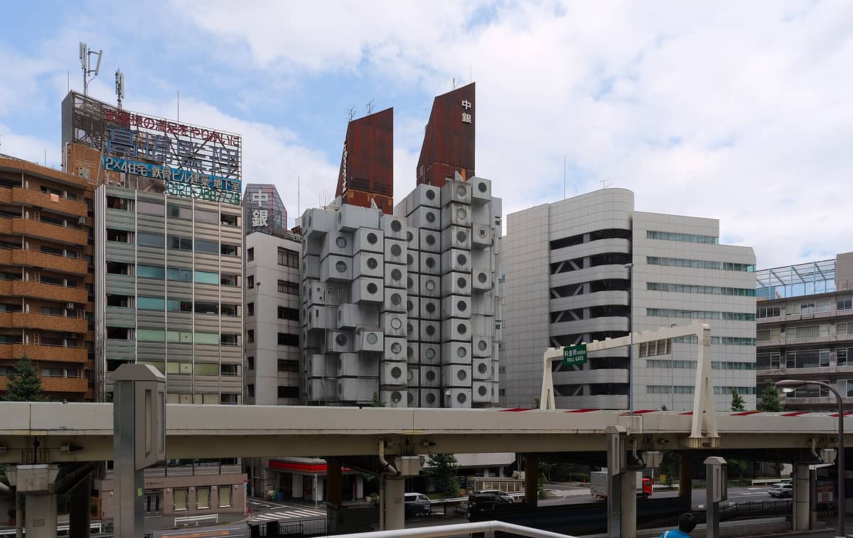 Unusual modular building with stacked cube units and round windows, surrounded by typical Tokyo city architecture and roadway in the foreground.