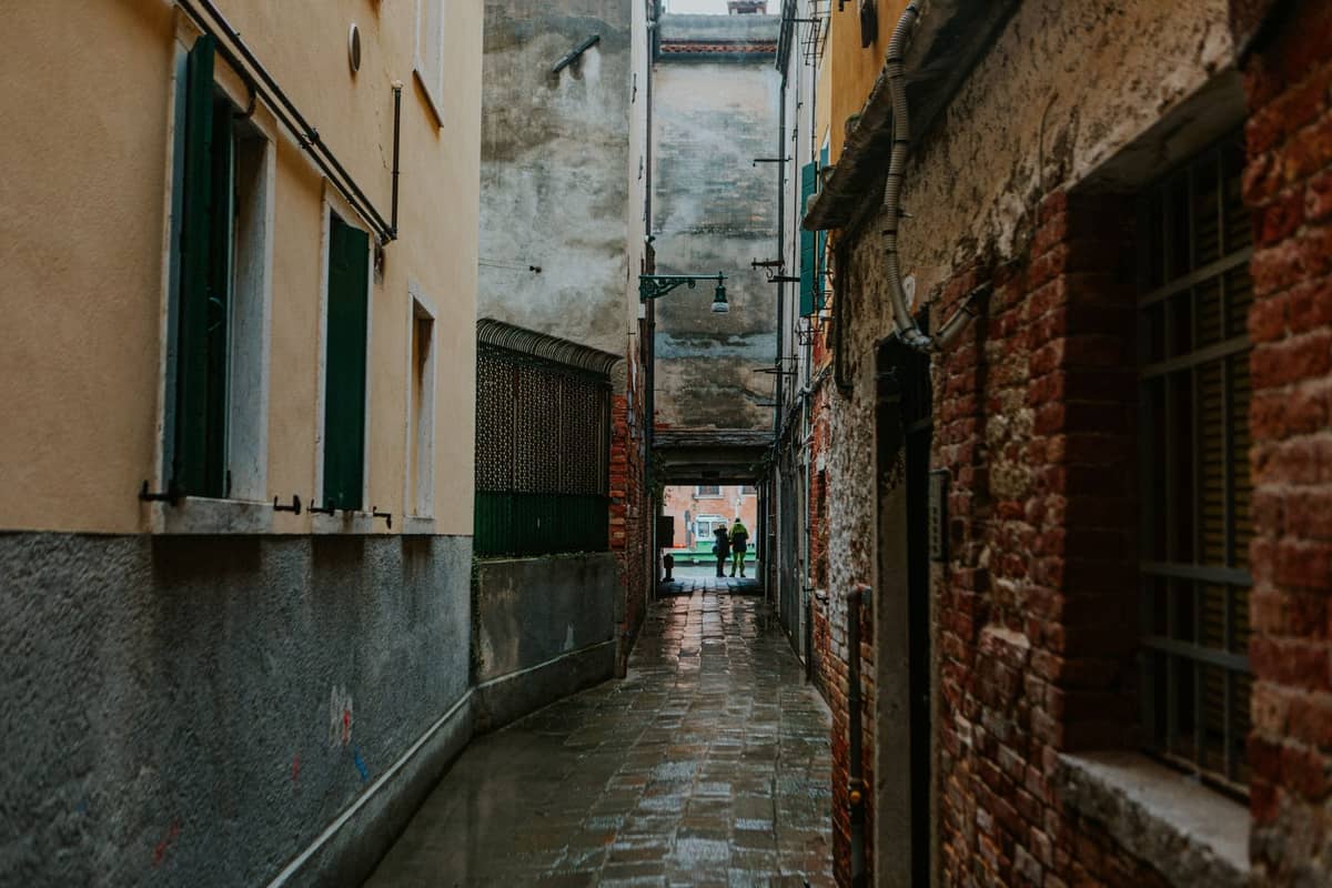 Enge Gasse in Venedig mit nassem Pflaster, verwitterten Mauern und zwei Silhouetten am Ende – ein versteckter Winkel fernab der Touristenmassen.