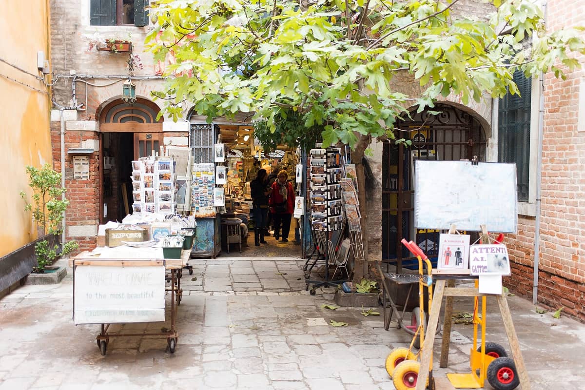 Außenhof-Eingang einer skurrilen Buchhandlung in Venedig mit Tischen, Bäumen, Postkarten und Menschen an der Tür.