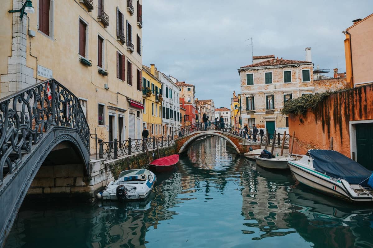 Ein ruhiger venezianischer Kanal mit Fußgängerbrücken, bunten Häusern, an den Seiten vertäuten Booten und Fußgängern im Hintergrund.