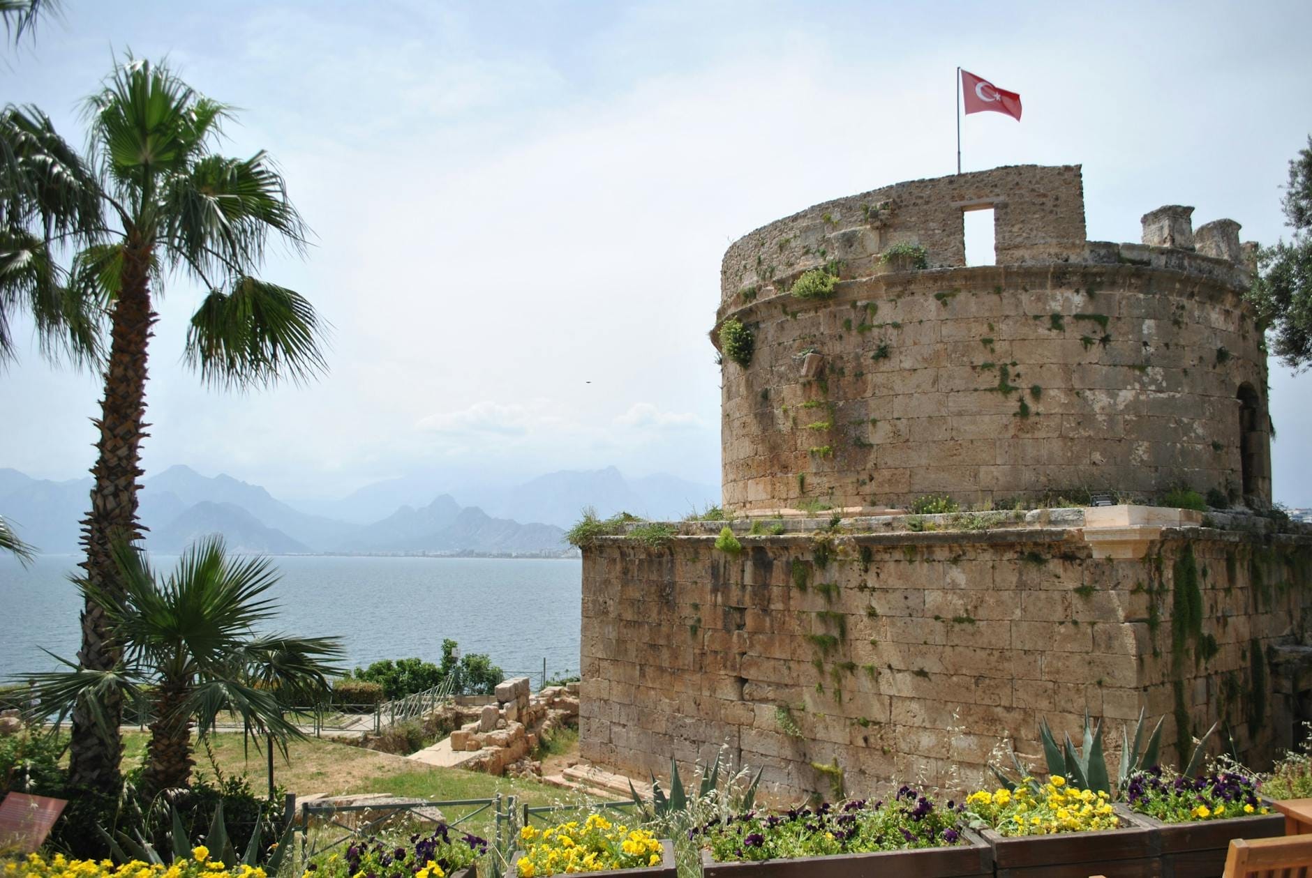 La Torre Hıdırlık, una struttura romana in pietra di forma circolare con una bandiera turca in cima, si erge sul mare con le montagne e le palme sullo sfondo.
