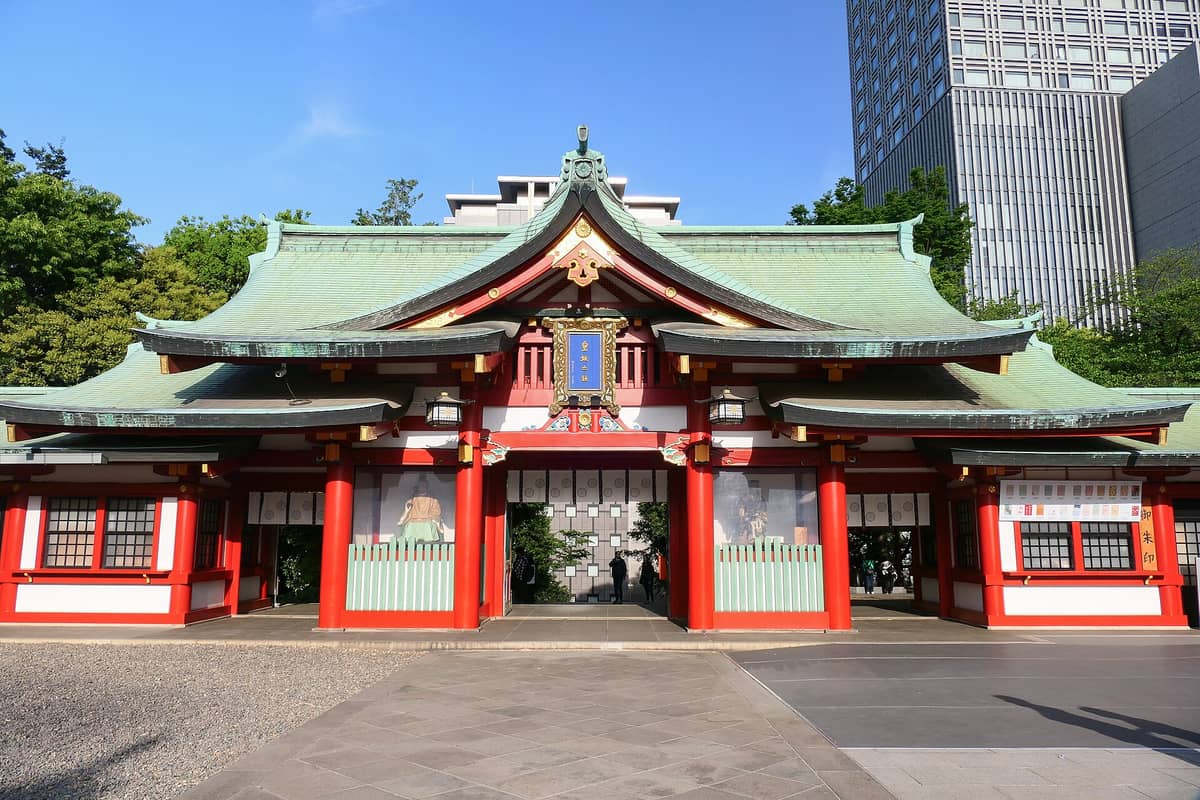 The main entrance gate of Hie Shrine in Tokyo, featuring vivid red pillars, green roofs, and modern buildings in the background on a bright sunny day.