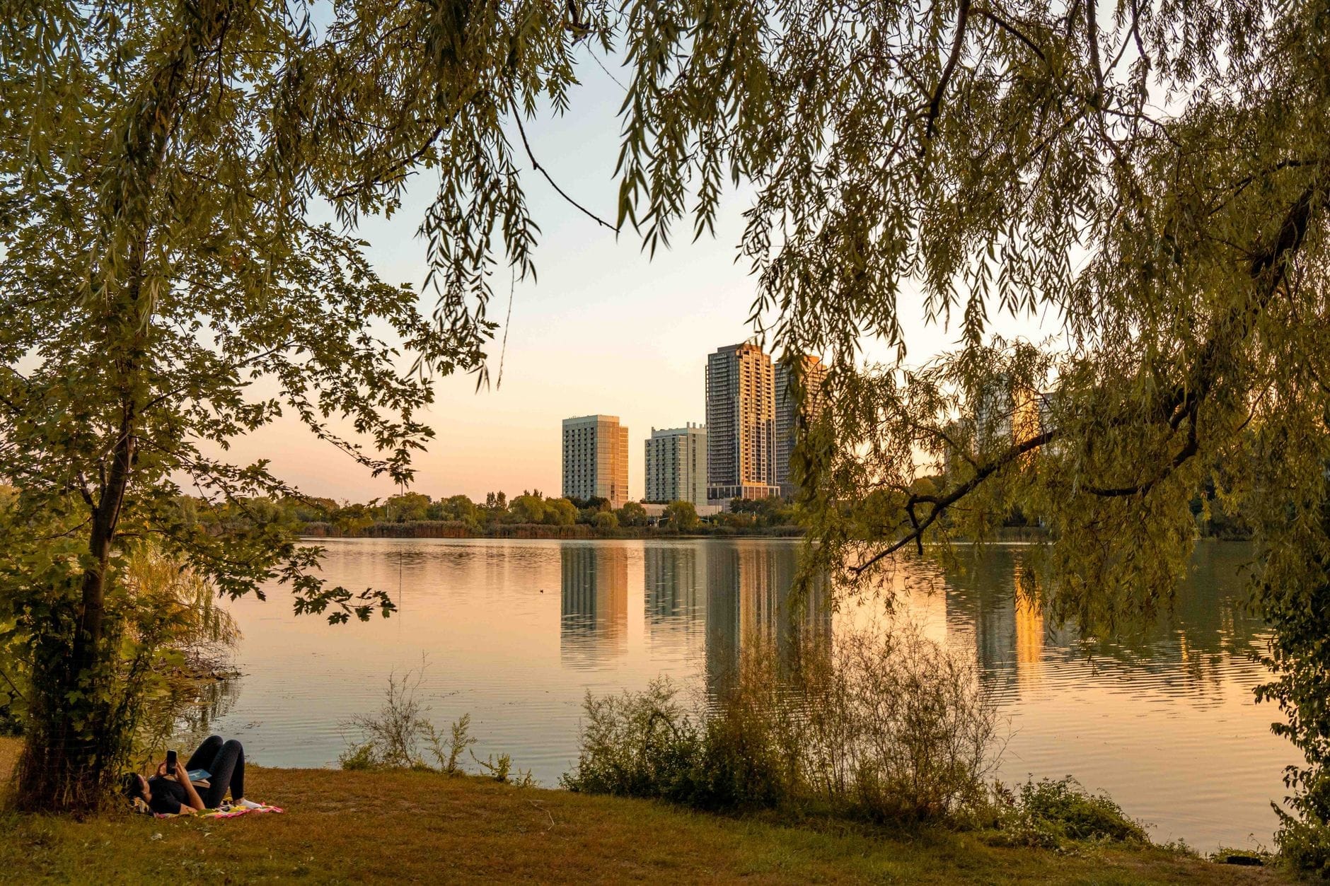 วิวสงบริมสระน้ำใน High Park ยามพระอาทิตย์ตก เงาไม้ล้อมกรอบ ตึกสูงสะท้อนบนผิวน้ำ ผู้คนปูเสื่อนั่งเล่นบนสนามหญ้า
