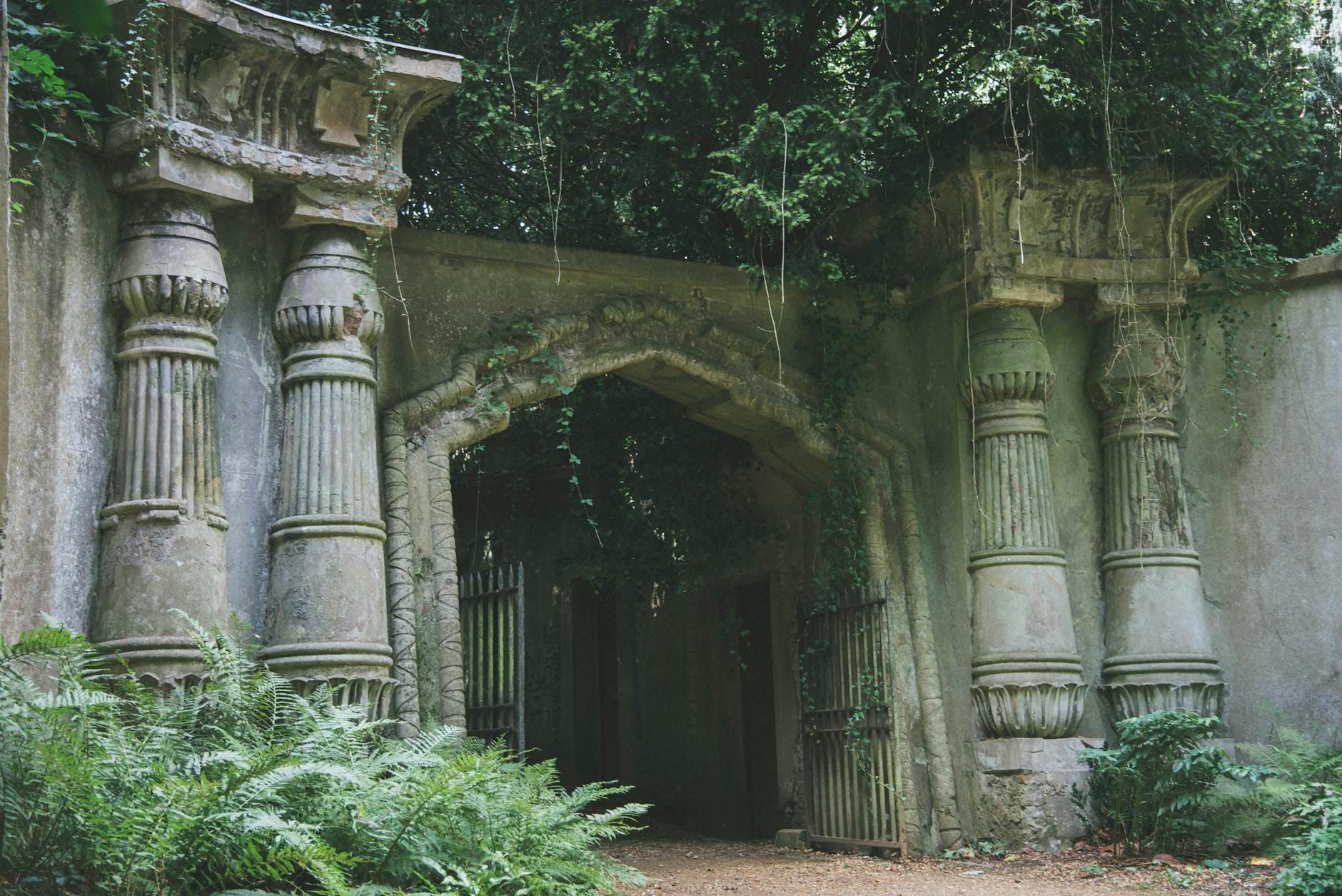 Antico arco di pietra vittoriano ricoperto di muschio, affiancato da imponenti pilastri e circondato da una vegetazione rigogliosa all'ingresso del cimitero di Highgate.