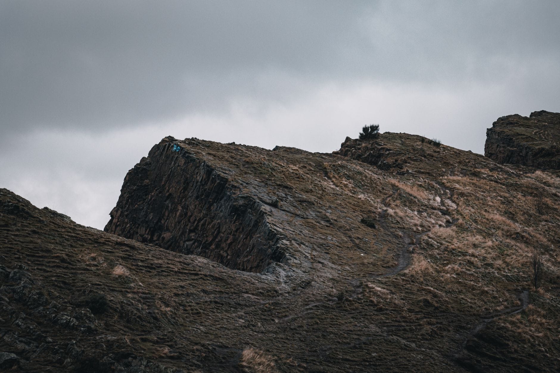 Dramatische Felsformationen und ein schmaler Wanderpfad auf einem windgepeitschten Hügel in Edinburgh unter einem bewölkten Himmel.