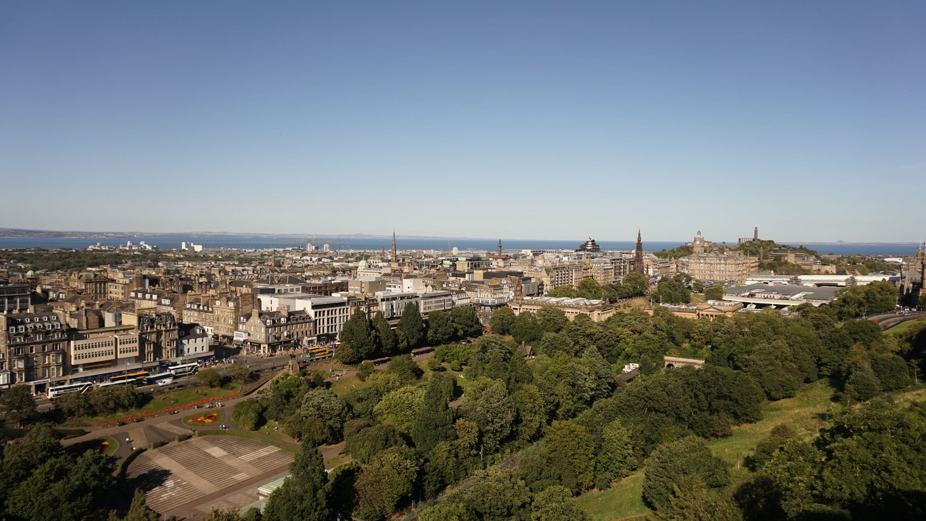 Weitläufiger Blick über die Princes Street Gardens mit üppigen grünen Rasenflächen, Bäumen und Sehenswürdigkeiten von Edinburgh unter einem klaren Himmel.