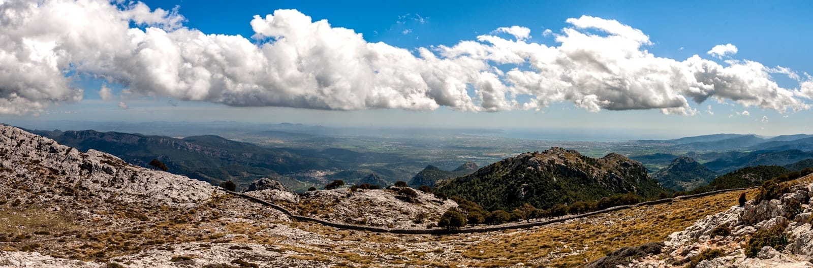 Vista panorâmica das Montanhas de Tramuntana com terreno rochoso, colinas onduladas, uma estrada sinuosa e nuvens dramáticas sob um céu azul intenso.