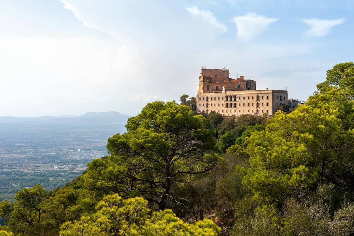 Historisches Klostergebäude auf einem Hügel, umgeben von Kiefern mit Blick auf die Ebenen von Mallorca, unter einem klaren Himmel.